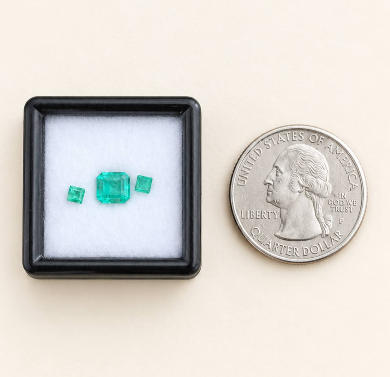 Cream background, black gem box, white cushion, and a U.S. quarter beside the box. One larger green center stone and two smaller green stones are inside the box. All three emeralds shown are included in the set.