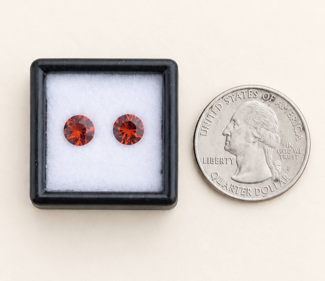 Cream background, black gem box, white cushion, and a U.S. quarter beside the box. Two round, faceted topaz stones are inside the box. The price is for the pair.