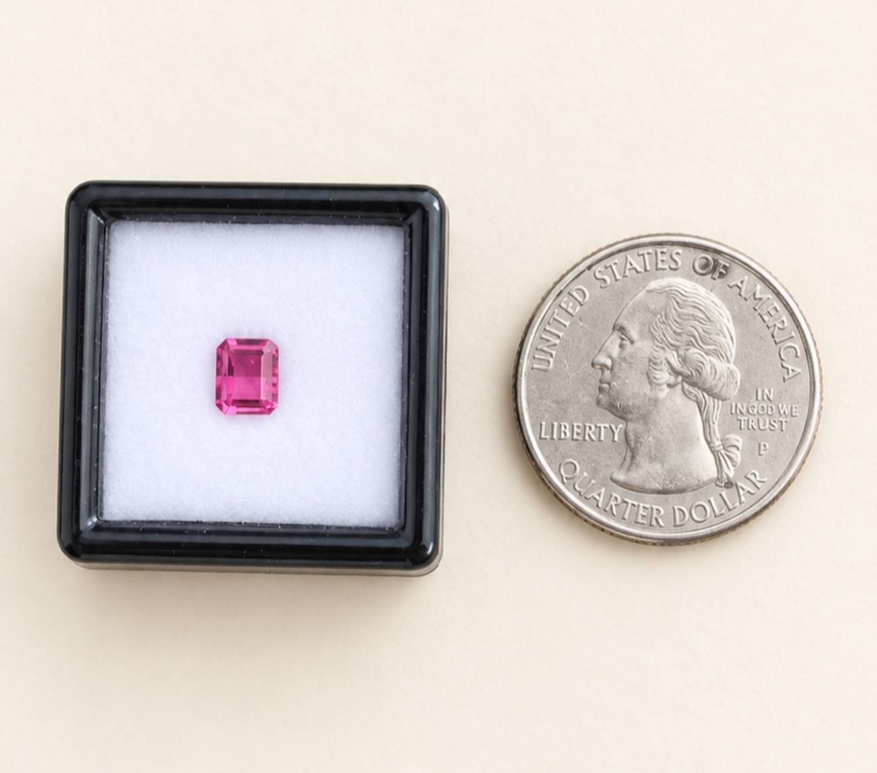 Cream background, black gem box, white cushion, and a U.S. quarter beside the box. One octagonal pink tourmaline is inside the box.